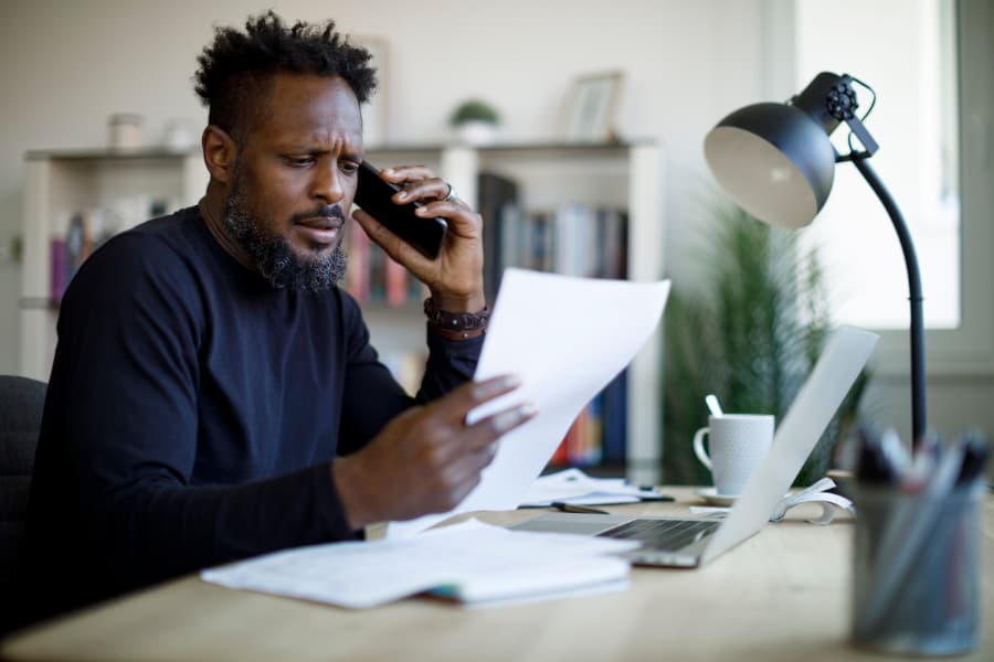 A confused looking man reading a paper and talking on the phone while sitting at a desk with a computer on it