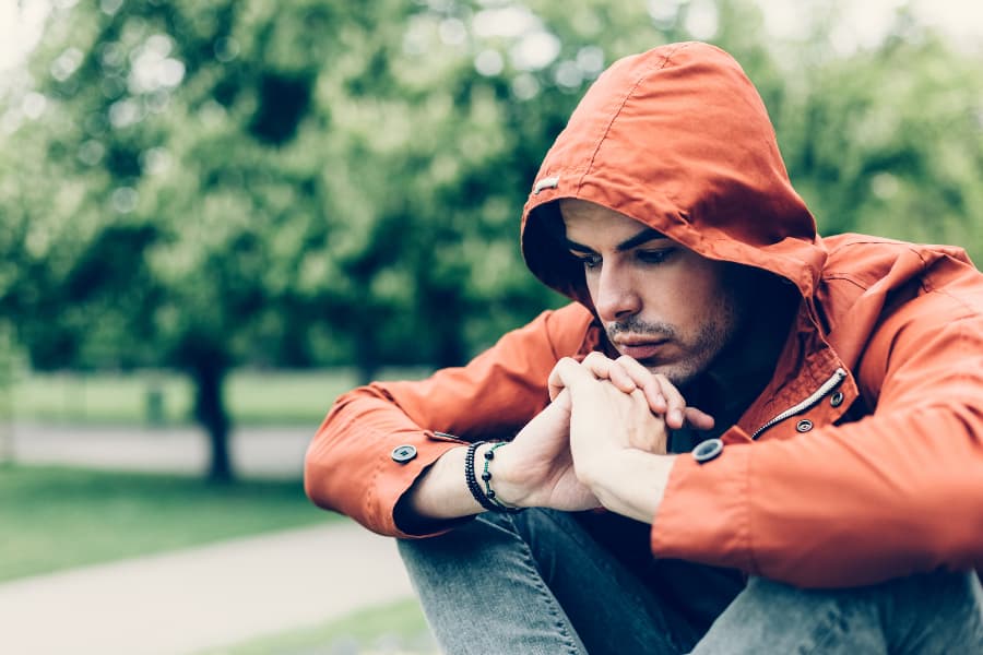 A depressed man wearing an orange jacket siting in a park with his hands clasped in front of him.