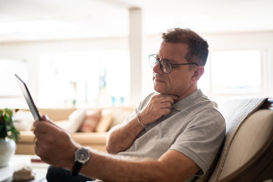 An older man looking at a tablet while touching his throat.