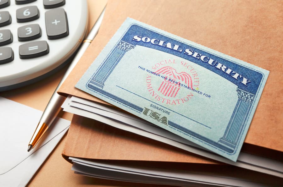 A Social Security card on top of a stack of file folders next to a calculator and pen.